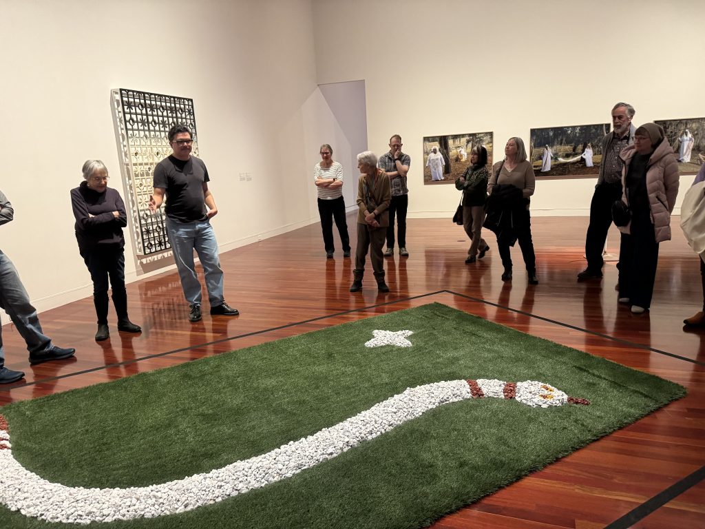 Members gather around an art piece composed of a piece of artificial turf with a snake formed on it with white and red rocks.
