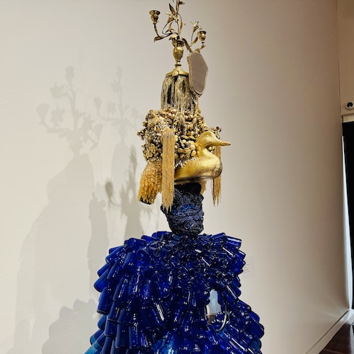 A sculpture made from found objects. The base is constructed of blue bottles and the top of gold items, including a duck, a hand mirror, and an ornate candlestick.