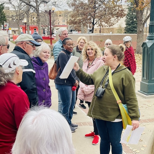 A woman in a green jacket speaks to a gathered tour group while holding up a sign