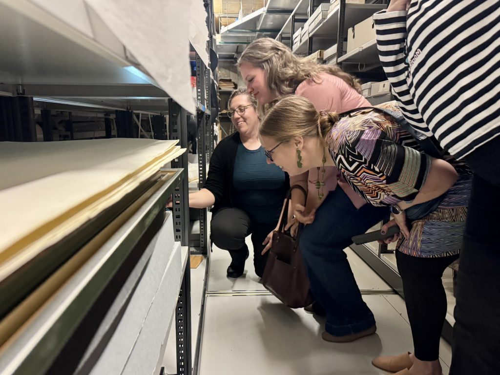 Four women bend over to look at some art pieces on a bottom shelf in storage