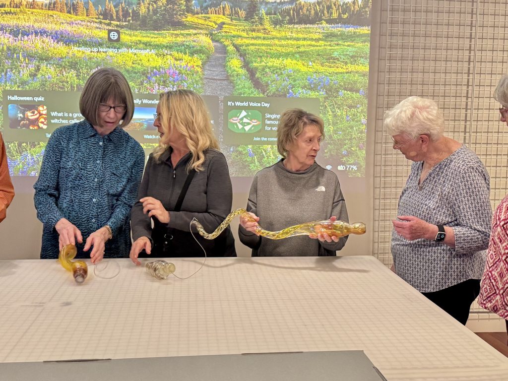 Four women hold blown glass pieces from a larger museum display around a large work table