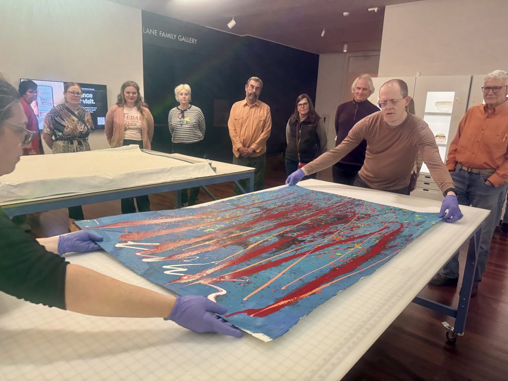 Two curators wearing gloves stretch out a large painting on a work table with museum members looking on
