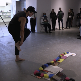 Josh T Franco performs Scriptorium con safos: Yard. He poses next to a display of books and magazines on the ground held down by large painted rocks.