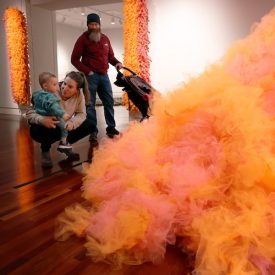 A woman squats with a toddler on her lap as they both look at an art piece made from heaps of orange gauze. A man with a baby stroller looks on.