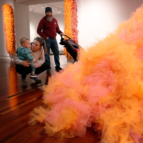 A woman squats with a toddler on her lap as they both look at an art piece made from heaps of orange gauze. A man with a baby stroller looks on.