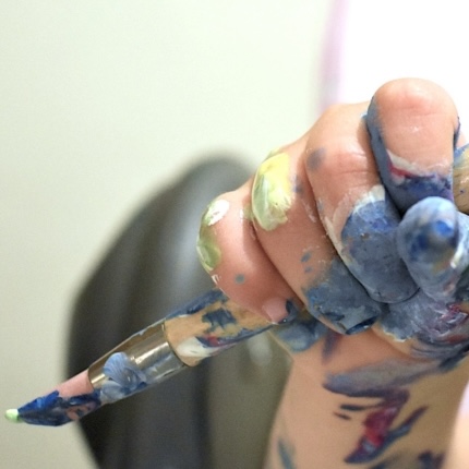 A toddler hand covered in paint holds a messy paintbrush