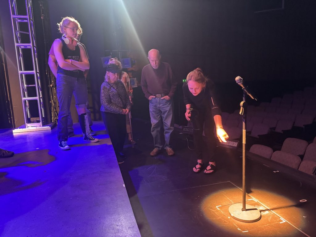 members stand on stage and look as a presenter points out a microphone in a spotlight where a platform is set to raise