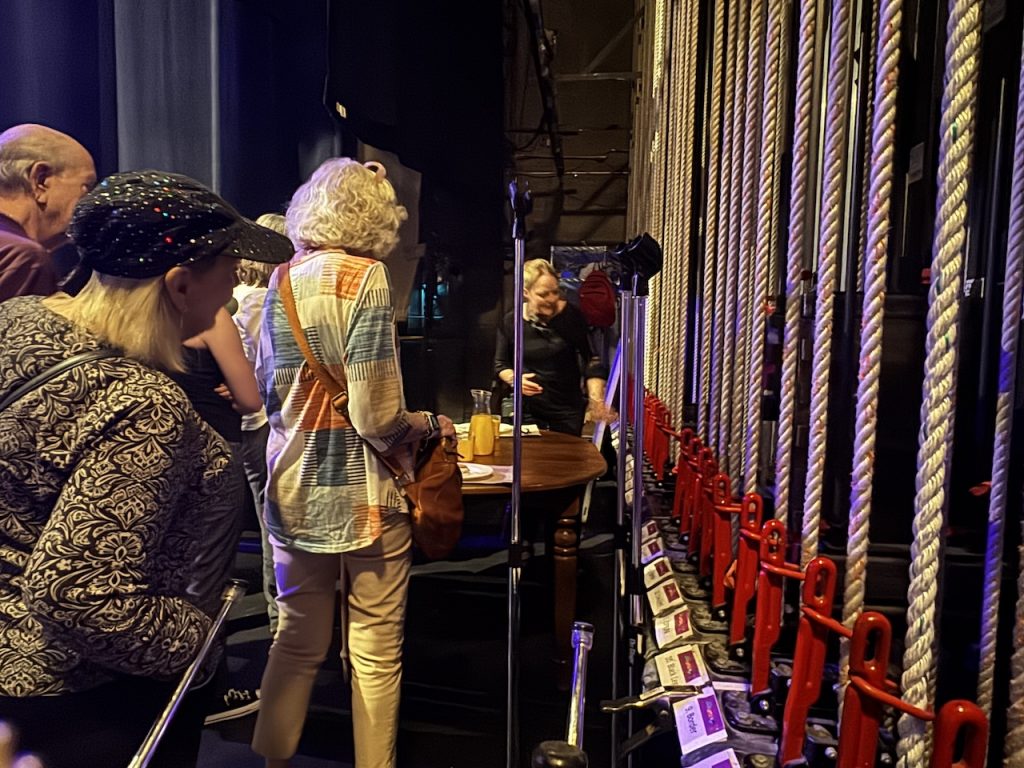 members stand backstage looking at a series of ropes set up for props and backdrops