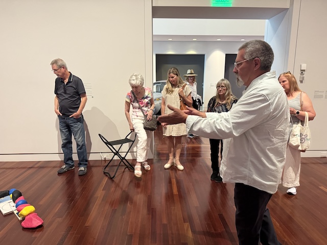 Man presents an art exhibit containing painted rocks on the ground to a group of gathered museum members
