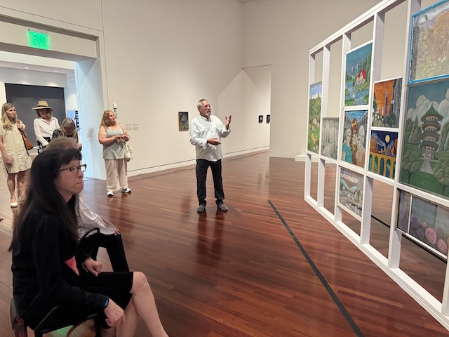 Man presents a wall of images in an exhibit to a gathered group of museum members