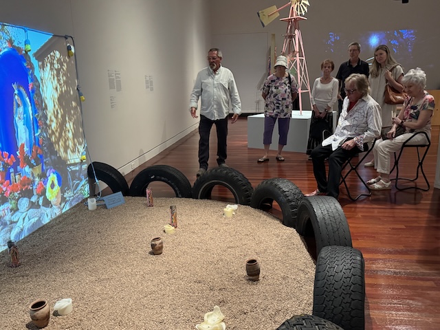 Group of museum members gathered in front of an art installation composed of gravel bordered by tires with a screen displaying an altar to the Virgin Mary. There are partially burned candles and ceramic containers on the gravel.