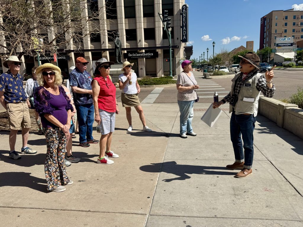 tour guide points to something out of frame for a group of members standing on the sidewalk downtown