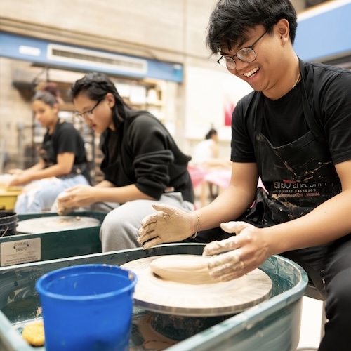 A young man in the foreground shapes clay on a potter's wheel while smiling. Two other students work with clay in the background.