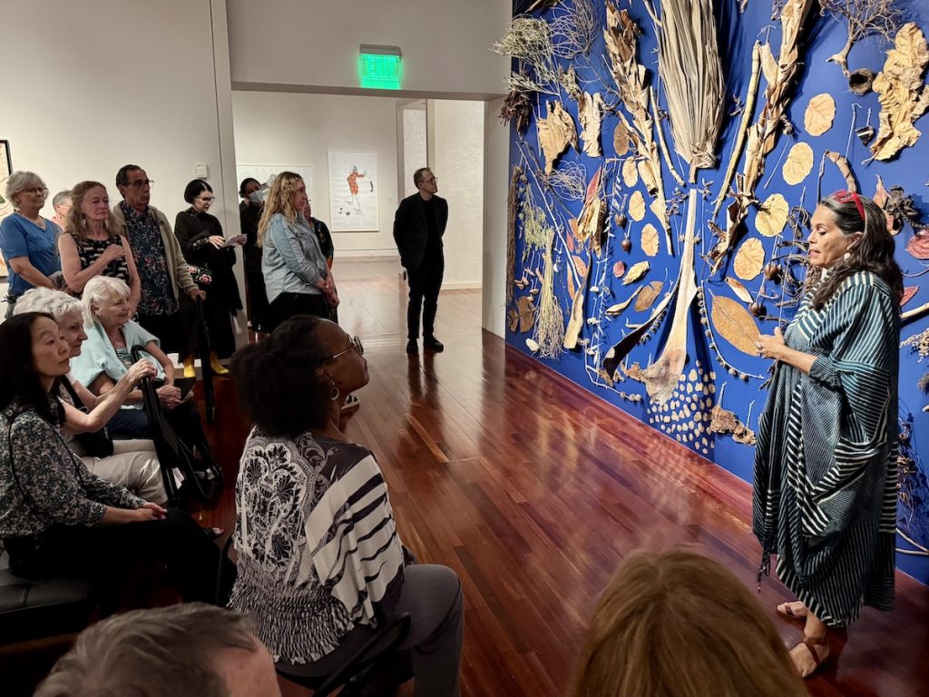 Members sit in a gallery and watch a presentation next to a large art installation on a blue wall