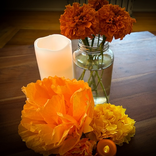 Paper marigolds sit in front of a lit candle next to a jar vase with marigolds.
